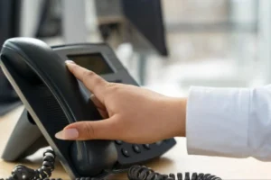 Business professional lifting a black desk phone receiver in office.